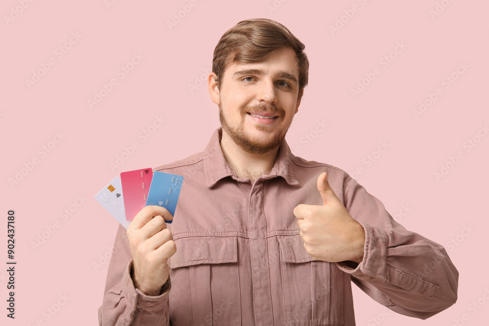 Smiling young man with credit cards showing thumb-up on pink background Smiling young man with credit cards showing thumb-up on pink background