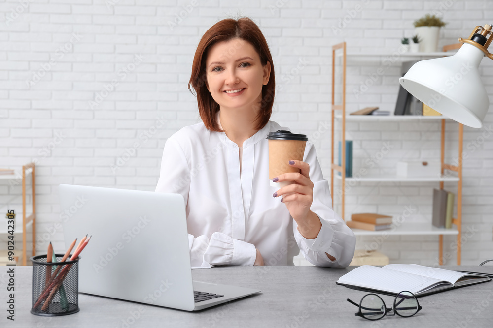 Young businesswoman with cup of coffee sitting at table with modern laptop in stylish office Young businesswoman with cup of coffee sitting at table with modern laptop in stylish office
