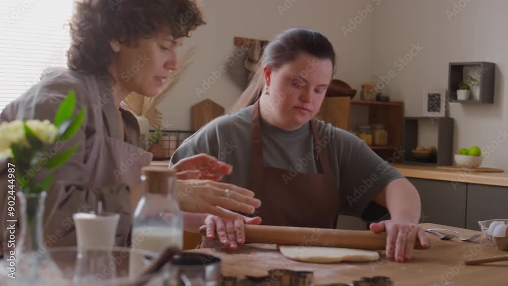 Female tutor teaching girl with Down syndrome to roll dough and sifting flour over rolling pin during process
