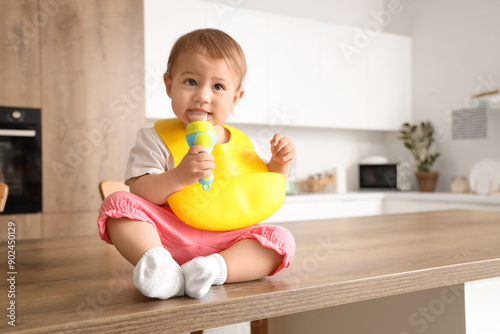 Cute little baby with bib and nibbler eating food on table in kitchen photo