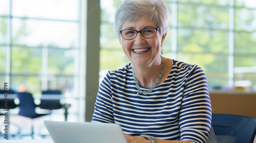 
An elderly woman with short gray hair, wearing glasses and a striped shirt. She is seated at a table with a laptop in front of her. The background reveals a well-lit office space with large windows