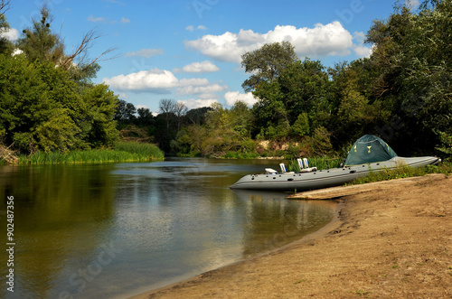 Moored inflatable boat sits peacefully on the river bank while trees form a serene backdrop, creating a piece of environmental art under a blue sky dotted with white clouds. photo