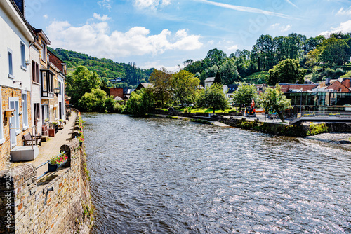 River Ourthe in village of La Roche-en-Ardenne, stone walls, houses, buildings and pedestrian path along river bank, mountains with green trees in misty background, sunny summer day in Belgium photo