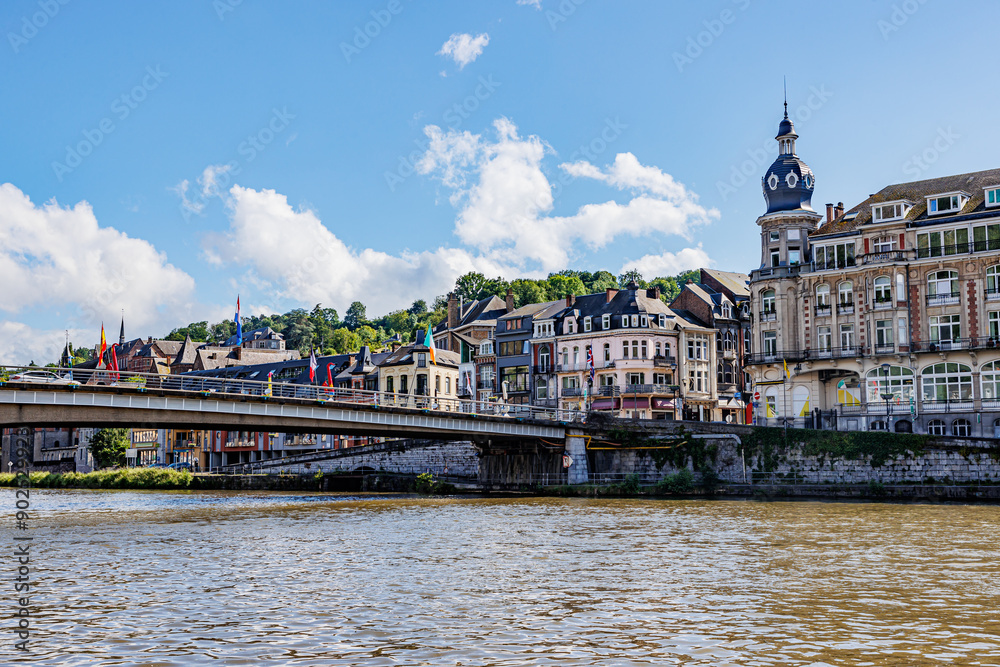 Fototapeta premium Cityscape of Dinant city, Meuse river, vehicular bridge with waving flags, cars driving, buildings and hills with leafy trees in background, sunny summer day in Namur province, Wallonia, Belgium