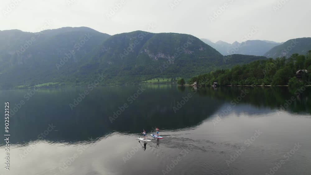 Drone shot over Lake Bohinj in Slovenia as a couple does stand up paddling (SUP) with mountains in the backdrop as the sun sets. Drone shot over Lake Bohinj in Slovenia as a couple does stand up paddling (SUP) with mountains in the backdrop as the sun sets.