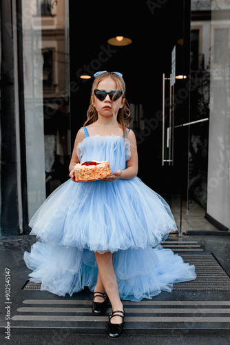 Elegant little girl in a lush elegant blue dress and sunglasses carries a birthday cake outdoor