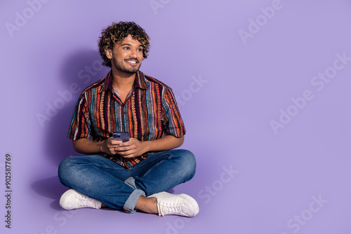 Full size photo of smart man dressed print shirt jeans sit on floor hold smartphone look empty space isolated on purple color background