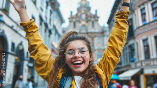 A happy young woman in a yellow jacket, glasses and ponytail raises her arms in joy on a rainy city street with European architecture. © COK House