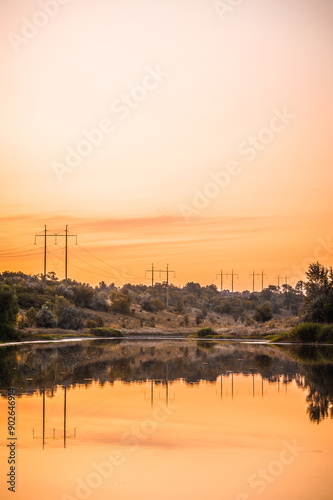 a calm lake with hills dotted with power poles in the background and an orange sunset sky in the background