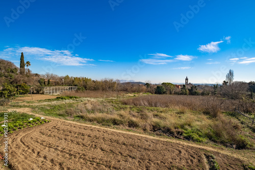 Expansive rural landscape with patches of farmland, distant trees, a clear blue sky with few clouds, and a hint of a tower in the distance.