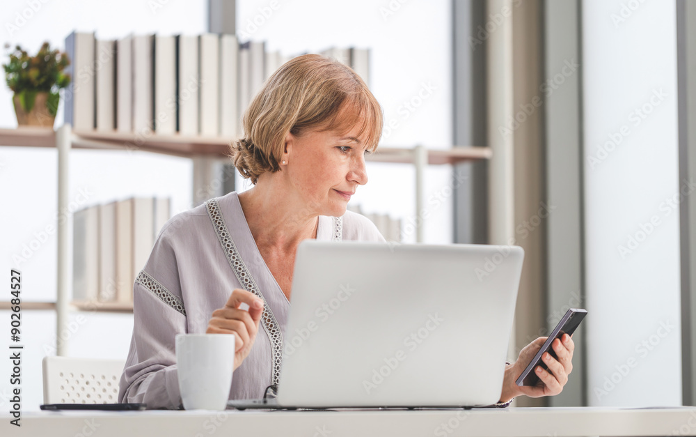 A middle-aged woman works at the table and looking at the smartphone, Woman using smartphone and laptop A middle-aged woman works at the table and looking at the smartphone, Woman using smartphone and laptop