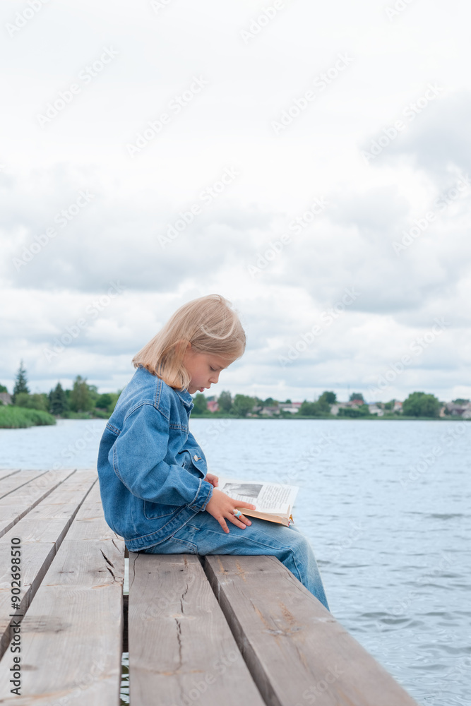 Fototapeta premium Girl reads a book while sitting on the pier. Education and learning concept. Vertical orientation.