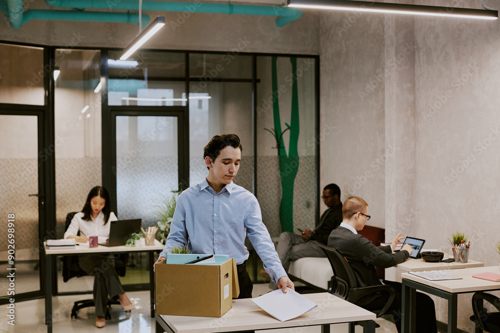 Young biracial man standing at desk in office of leading company getting ready for first workday, taking documents and stuff out of box Young biracial man standing at desk in office of leading company getting ready for first workday, taking documents and stuff out of box