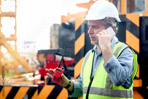 Senior worker engineer foreman with safety vest and helmet talking on mobile phone while working at construction site. Mature elderly architect works at ground level of building workplace.