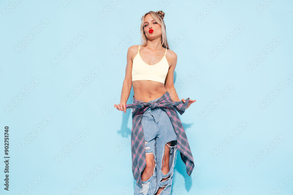 Young beautiful smiling female in trendy summer jeans and top clothes, plaid shirt tied on waist. Carefree woman posing near blue wall in studio. Sexy positive model. Red lips
