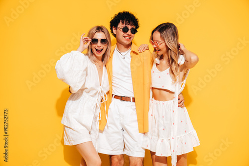 Group of young three stylish friends posing in studio. Fashion man and two cute female dressed in casual summer clothes. Smiling models having fun. Cheerful women and guy near yellow wall, sunglasses © halayalex