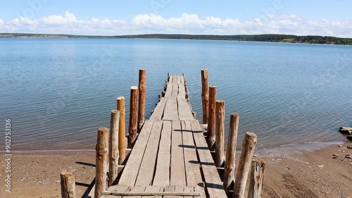 Long wooden footway extends across calm blue river, with a sandy shore on either side and beautiful blue sky overhead.
