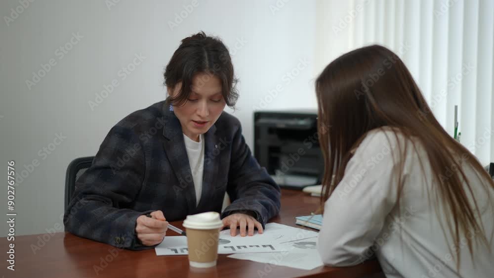 Two business women discussing a chart while sitting at a table in an office. A woman manager points with a pen at a chart on paper, lying on the table. A plastic cup of coffee stands on the desktop Two business women discussing a chart while sitting at a table in an office. A woman manager points with a pen at a chart on paper, lying on the table. A plastic cup of coffee stands on the desktop