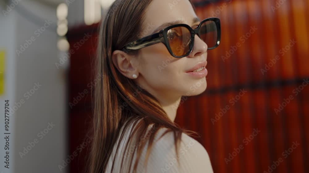 A fashionable young woman with long brown hair wears large sunglasses, basking in warm sunlight with a thoughtful look, against a vibrant urban backdrop. Stylish Young Woman in Sunglasses Enjoying A fashionable young woman with long brown hair wears large sunglasses, basking in warm sunlight with a thoughtful look, against a vibrant urban backdrop. Stylish Young Woman in Sunglasses Enjoying