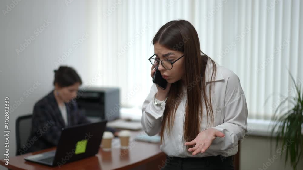 In the foreground, a girl is holding a cell phone and talking emotionally on it while standing in the office. In the background in the defocus, a woman is working on a laptop sitting at her desk In the foreground, a girl is holding a cell phone and talking emotionally on it while standing in the office. In the background in the defocus, a woman is working on a laptop sitting at her desk