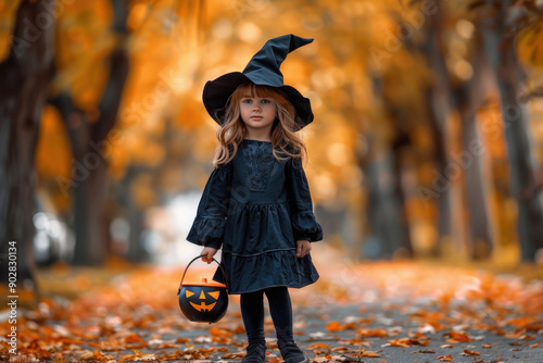 Child dressed as a witch for Halloween holding a pumpkin bucket standing on a road in an autumnal forest photo