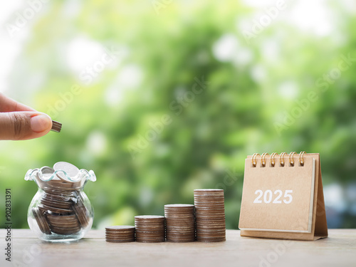 Close up hand putting coin into glass bottle, 2025 desk calendar and stack of coins. The concept of saving money, Financial, Investment and Business growing in year 2025.