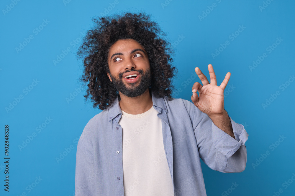 Young male with curly hair and playful smile gestures an 'OK' sign with his right hand, radiating positivity and confidence. He stands against a lively blue backdrop, embodying a cheerful vibe. Young male with curly hair and playful smile gestures an 'OK' sign with his right hand, radiating positivity and confidence. He stands against a lively blue backdrop, embodying a cheerful vibe.