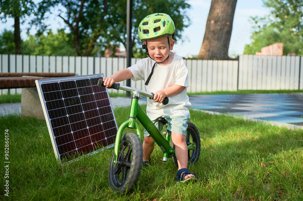 Young boy sits on green balance bike next to solar panel on grass. Kid wearing green helmet. Background features fence, trees, and bench. Concept of green energy generation. Young boy sits on green balance bike next to solar panel on grass. Kid wearing green helmet. Background features fence, trees, and bench. Concept of green energy generation.