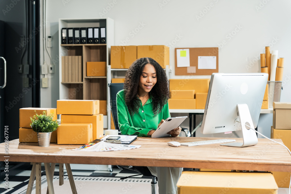 An African American woman with an afro works at a wooden desk, equipped with a laptop, desktop computer, and parcel cardboard boxes, focusing on SME logistics, inventory, and online orders. An African American woman with an afro works at a wooden desk, equipped with a laptop, desktop computer, and parcel cardboard boxes, focusing on SME logistics, inventory, and online orders.