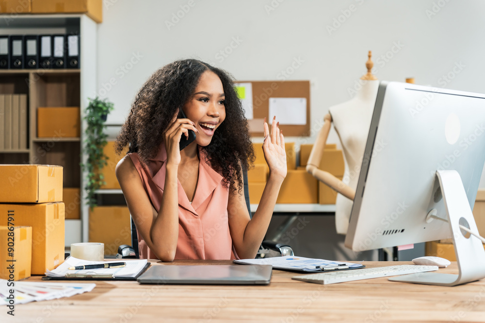 Obraz premium An African American woman with an afro works at a wooden desk, equipped with a laptop, desktop computer, and parcel cardboard boxes, focusing on SME logistics, inventory, and online orders.