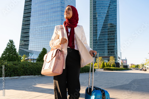 Beautiful arab businesswoman wearing hijab and elegant business suit portrait in the city - Middle eastern adult female on business travel walking with baggage