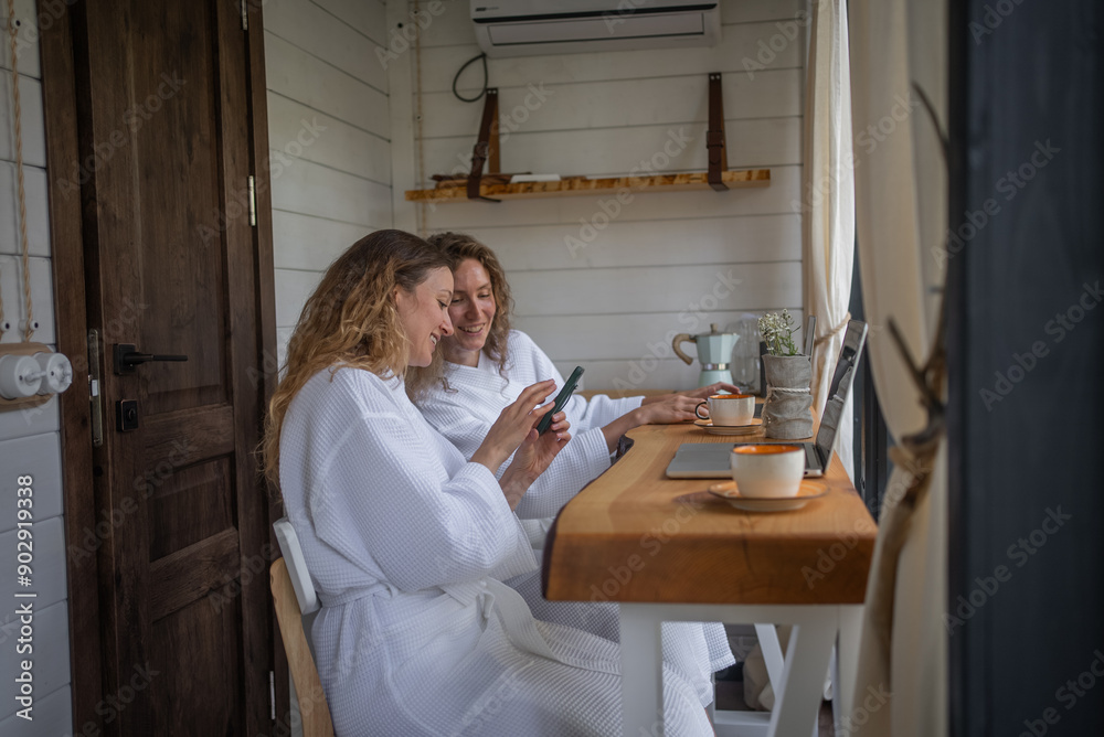 Woman girlfriends shows her friend a funny news or photo on her smartphone while sitting at her laptop desk. Girls work online remotely outdoors. Woman girlfriends shows her friend a funny news or photo on her smartphone while sitting at her laptop desk. Girls work online remotely outdoors.