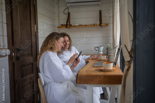 Woman girlfriends shows her friend a funny news or photo on her smartphone while sitting at her laptop desk. Girls work online remotely outdoors.