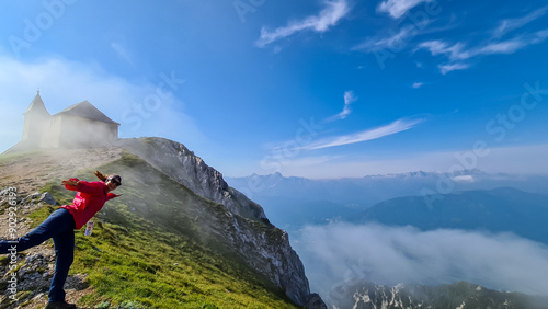 Woman walking on scenic hiking trail with panoramic view of chapel Maria am Stein near Dobratsch, Villacher Alps, Carinthia, Austria, Europe. Looking at Julian and Karawanks mountain range. Wanderlust photo