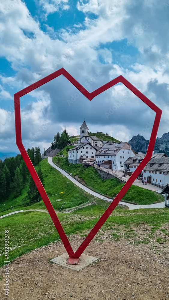 Heart shaped landmark with view of alpine village of Monte Santo di Lussari, Julian Alps, Tarvisio, Friuli Venezia Giulia, Italy. Looking at majestic mountain ridges. Family trip on cloudy summer day Heart shaped landmark with view of alpine village of Monte Santo di Lussari, Julian Alps, Tarvisio, Friuli Venezia Giulia, Italy. Looking at majestic mountain ridges. Family trip on cloudy summer day
