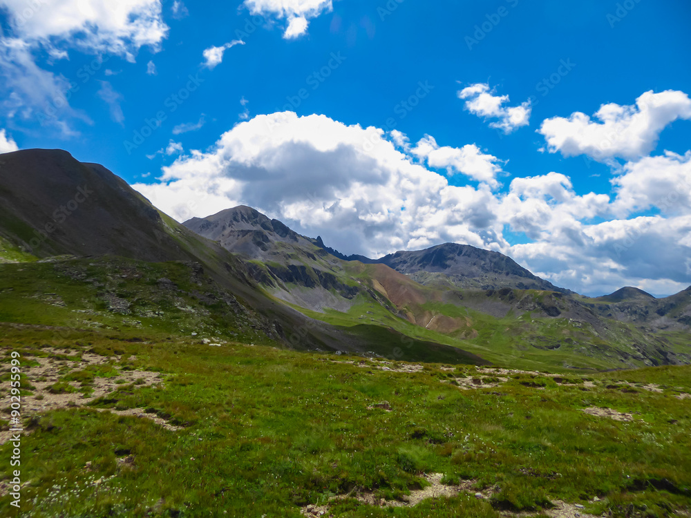 Fototapeta premium Alpine green pasture with panoramic view of mountains and hills in valley Valle Stura in Colle Del Puriac, Piemonte, Italy, Europe. Hiking tour in Italian Alps in summer. Peaceful serene atmosphere