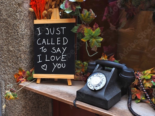A romantic setup with a vintage black dial rotary phone placed next to a blackboard. On the blackboard, a heartfelt message is handwritten in chalk, stating I just called to say I LOVE you