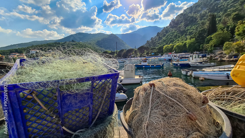 A close up on fishing nets and boats parked along the shores of Medveja, Croatia. There are plenty of buoys in the water. The Mediterranean Sea is calm. Stony beach and lusch green mountains behind photo