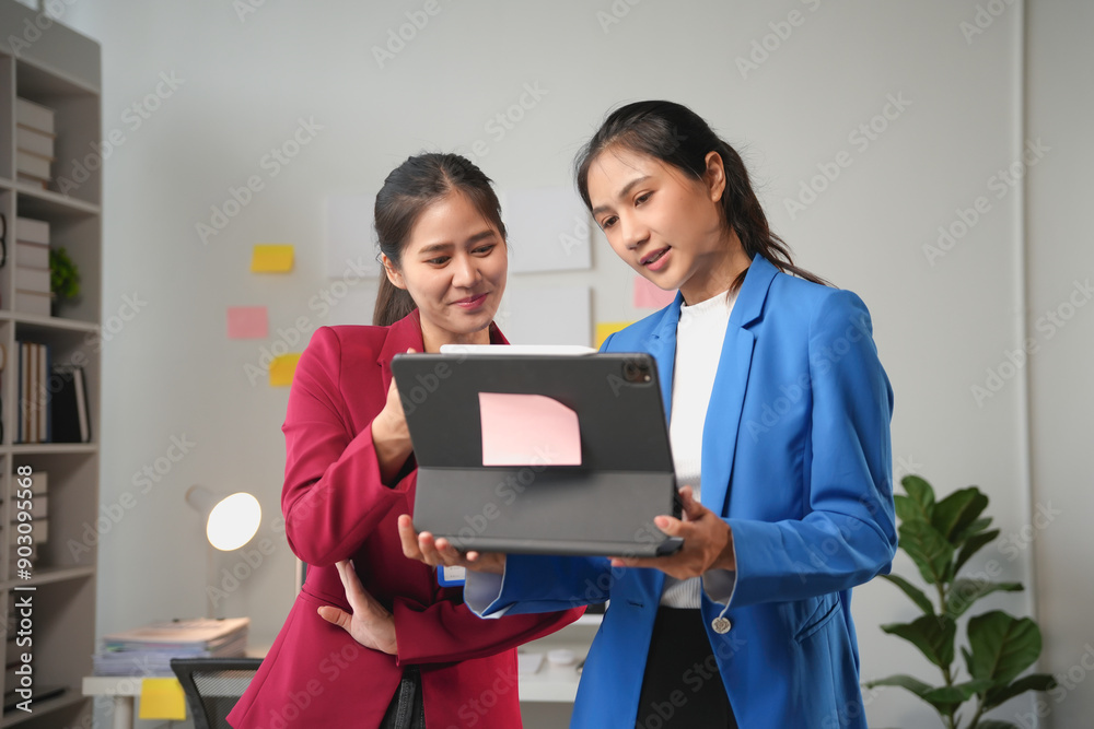Two businesswomen are standing together in their office, reviewing work on a digital tablet Two businesswomen are standing together in their office, reviewing work on a digital tablet