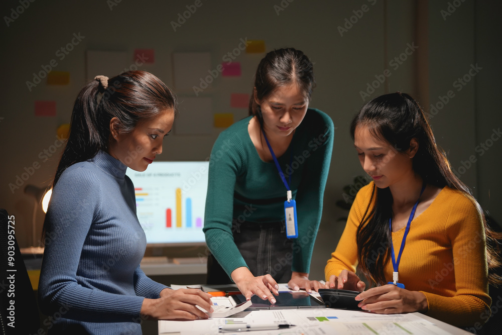 Three businesswomen are working late in the office, focused on a digital tablet and discussing a project Three businesswomen are working late in the office, focused on a digital tablet and discussing a project