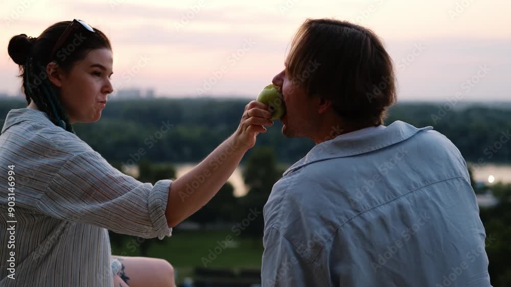 A young pretty Caucasian couple is sitting at sunset during a date in the park, drinking wine and eating a green apple. A girl with blue dreadlocks with young man in a stylish blue shirt A young pretty Caucasian couple is sitting at sunset during a date in the park, drinking wine and eating a green apple. A girl with blue dreadlocks with young man in a stylish blue shirt