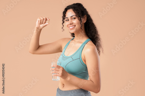 Young African-American woman taking fish oil on beige background