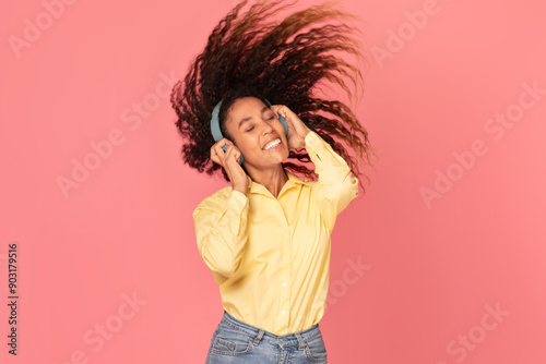 African American woman with curly brown hair wears headphones and dances while smiling, enjoying the music. She is wearing a yellow shirt and blue jeans and is posed in front of a pink background.