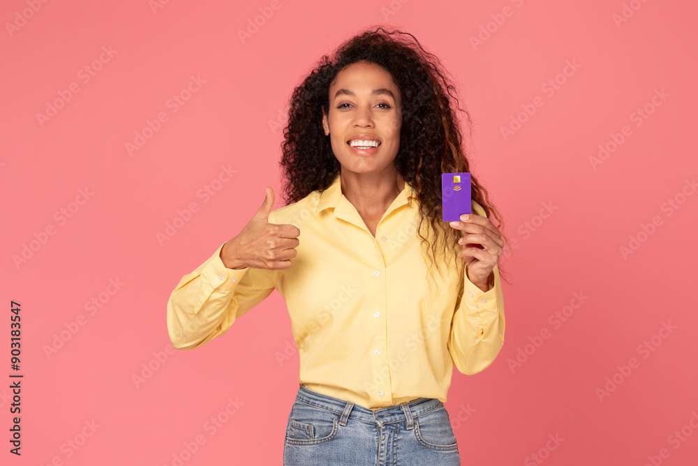 African American young woman with curly hair is smiling and giving a thumbs up while holding a purple credit card in her other hand. The background is a solid pink. African American young woman with curly hair is smiling and giving a thumbs up while holding a purple credit card in her other hand. The background is a solid pink.