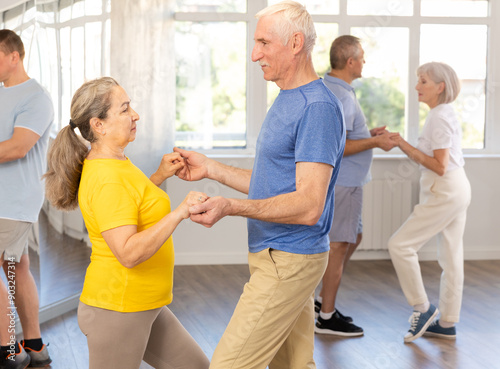 Positive senior man dancing playful Latin dance bachata with female partner during group class in choreography studio. Active lifestyle of older generation