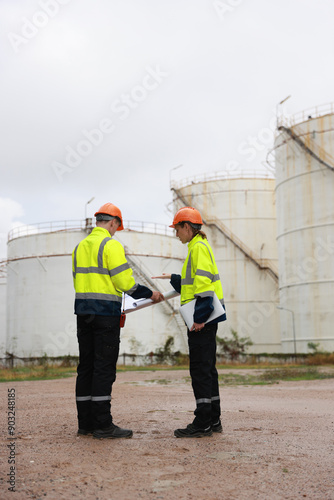 The senior manager female engineer and her male colleague are confidently working together at an oil storage tank field with a blueprint, utilizing a tablet to effectively track the progress of the pr photo