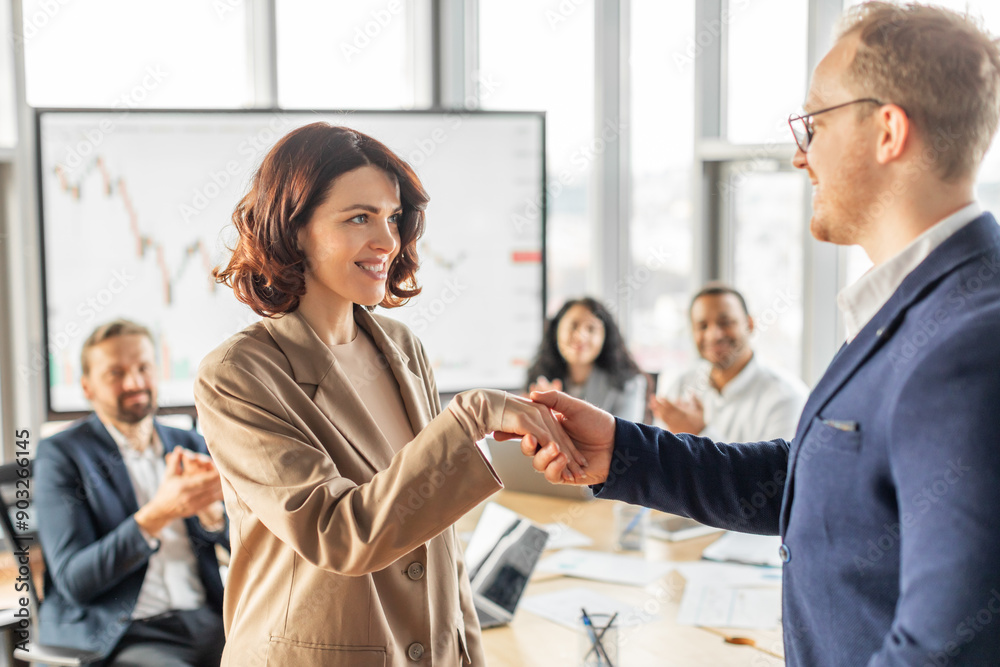 A businesswoman in a beige blazer shakes hands with a male colleague in a blue suit, smiling broadly. The woman is standing, her colleague is seated, and they are both in a modern office setting A businesswoman in a beige blazer shakes hands with a male colleague in a blue suit, smiling broadly. The woman is standing, her colleague is seated, and they are both in a modern office setting