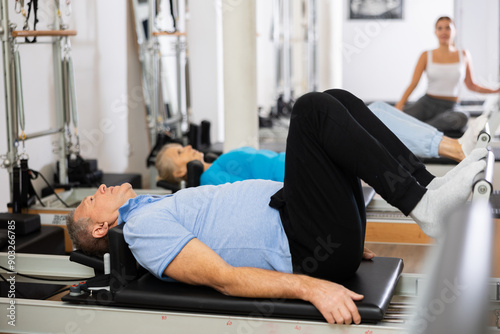 Concentrated old man doing Pilates exercises for lower back while lying on reformer bed in rehabilitation center