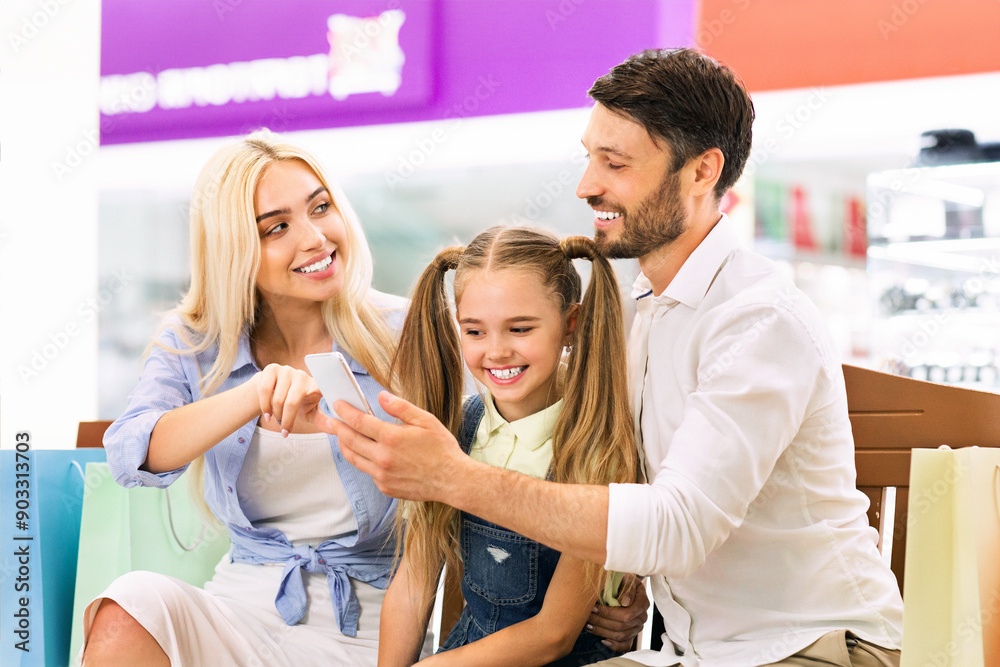 A young family, a father, a mother, and their young daughter, are sitting together in a shopping mall. They are looking at a smartphone together and smiling A young family, a father, a mother, and their young daughter, are sitting together in a shopping mall. They are looking at a smartphone together and smiling