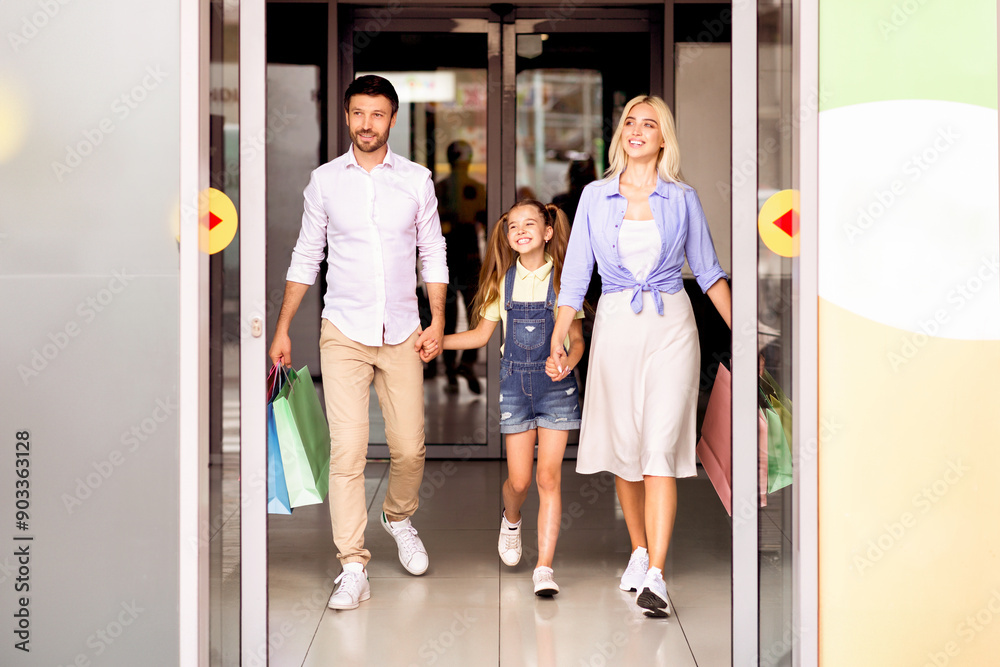 Fototapeta premium A family, consisting of a father, a mother, and a young daughter, walks out of a shopping mall, carrying shopping bags. The parents are holding hands with their daughter, and they are all smiling